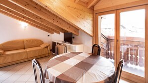 Ein Zimmer mit Holzbalkendecke, einer Couch, einem Tisch mit Stühlen, einem Fernseher und Blick durch eine Glastür.