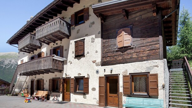A traditional chalet with wooden balconies, stone walls, and a staircase leading to the upper floors.