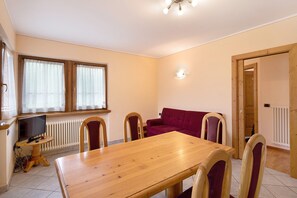 Un comedor con una mesa y sillas de madera, un televisor sobre una mesita, un sofá rojo y una ventana con cortinas.