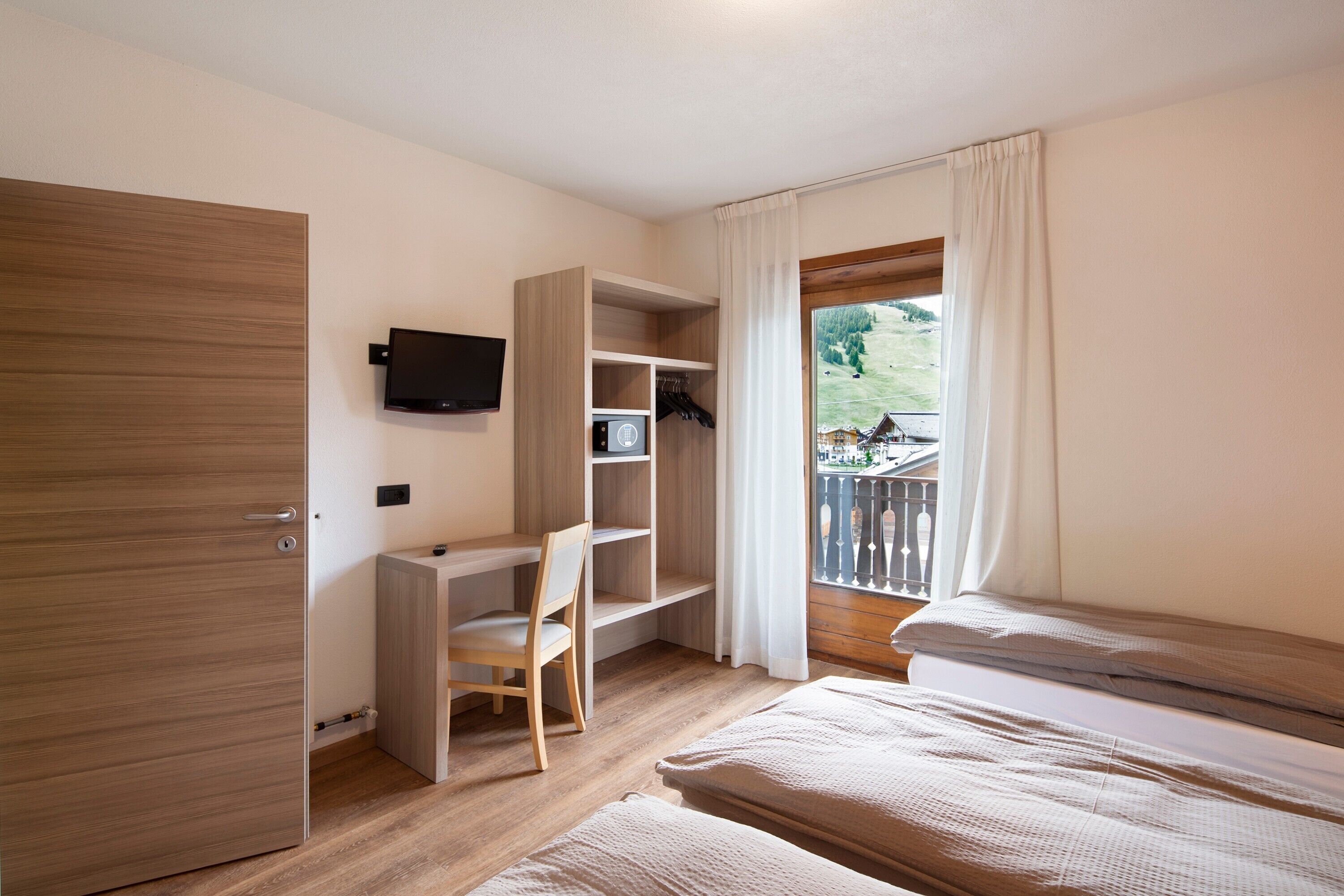 Ein Schlafzimmer mit Holztür, Schreibtisch mit Stuhl, Fernseher, Bett und einem Balkon mit Blick auf eine grüne Landschaft.