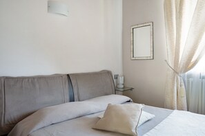 A bedroom with a bed, a bedside table with a lamp, a mirror on the wall, and a window with curtains.