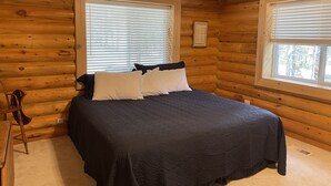 Un dormitorio de cabaña de troncos con una cama, dos ventanas con persianas, un cuadro enmarcado en la pared y una pequeña mesa con sillas.