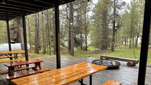 Una terraza de madera al aire libre con mesas de picnic y un hogar para fogata, rodeada de árboles.
