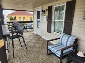 Une vĂ©randa couverte avec un plancher en bois, une table Ă manger avec des chaises, un canapĂ© avec des coussins Ă rayures et une fenĂȘtre avec des volets.