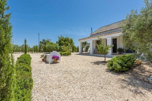 Un bâtiment blanc avec un toit en tuiles, entouré de gravier, d’arbres et d’un cactus.