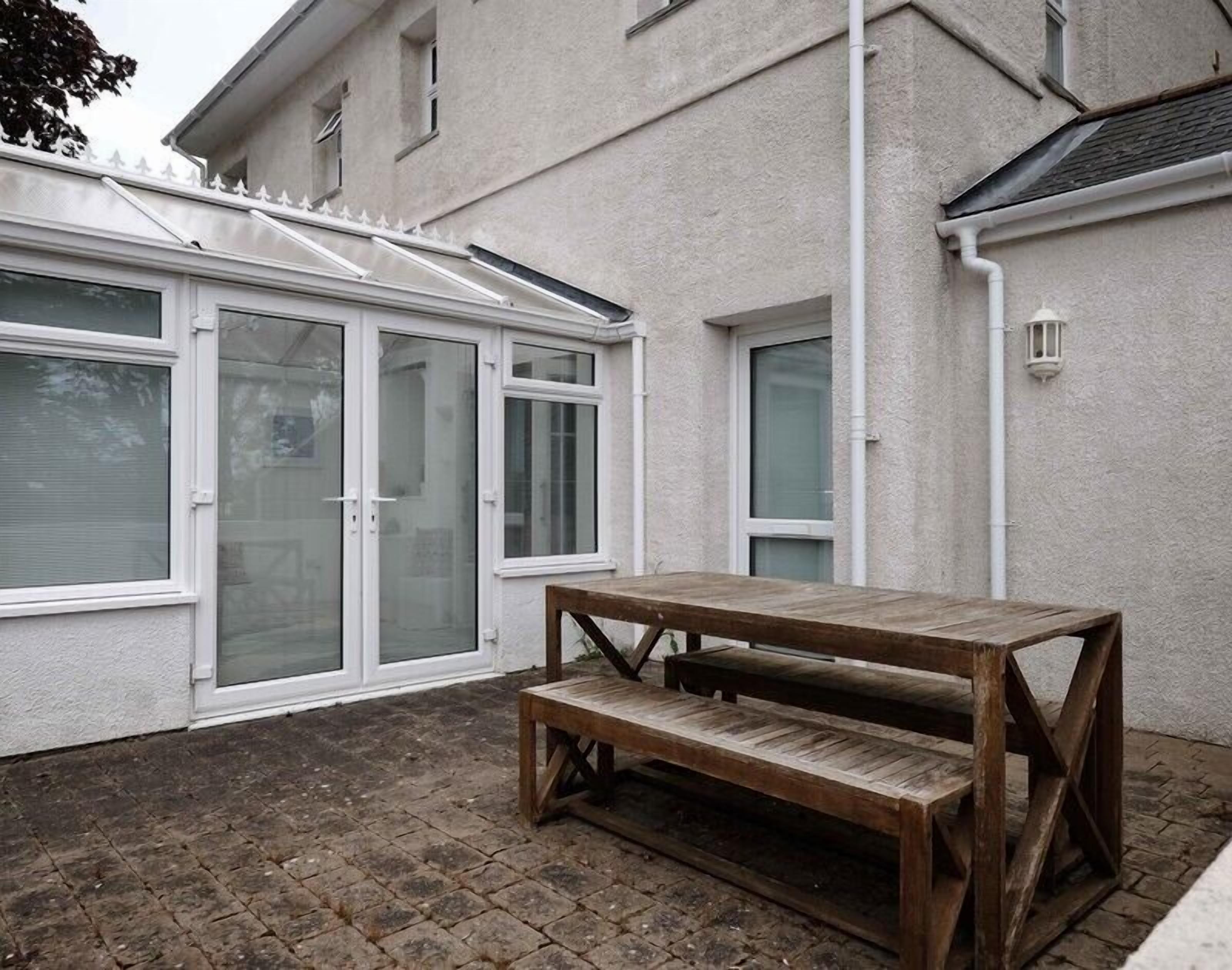 A patio with a wooden table and bench, a glass door entrance, and a white exterior wall.