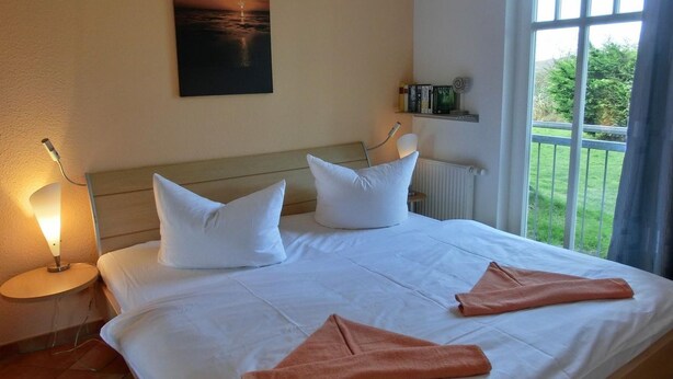 Habitación de hotel con cama, almohadas blancas, un cuadro de atardecer, ventanal con vistas a follaje y una estantería con libros y una lámpara.