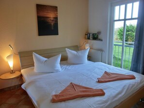 A hotel room with a bed, white pillows, a painting of a sunset, a window with a view of greenery, and a shelf with books and a lamp.