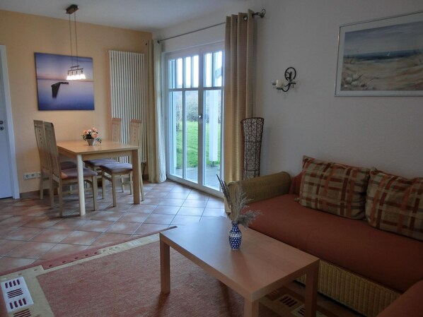 A living room with a sofa, a coffee table, a dining table with chairs, a painting, and a view through the glass door.
