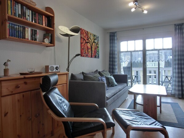 A modern living room with a sofa, chairs, a coffee table, a bookshelf, a painting, and a window with a view of buildings.