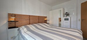 Ein Schlafzimmer mit einem Lattenrost-Kopfteil, einer gestreiften Bettdecke, Nachttischlampen, einem weißen Kleiderschrank und einem kleinen Topfpflanzen.