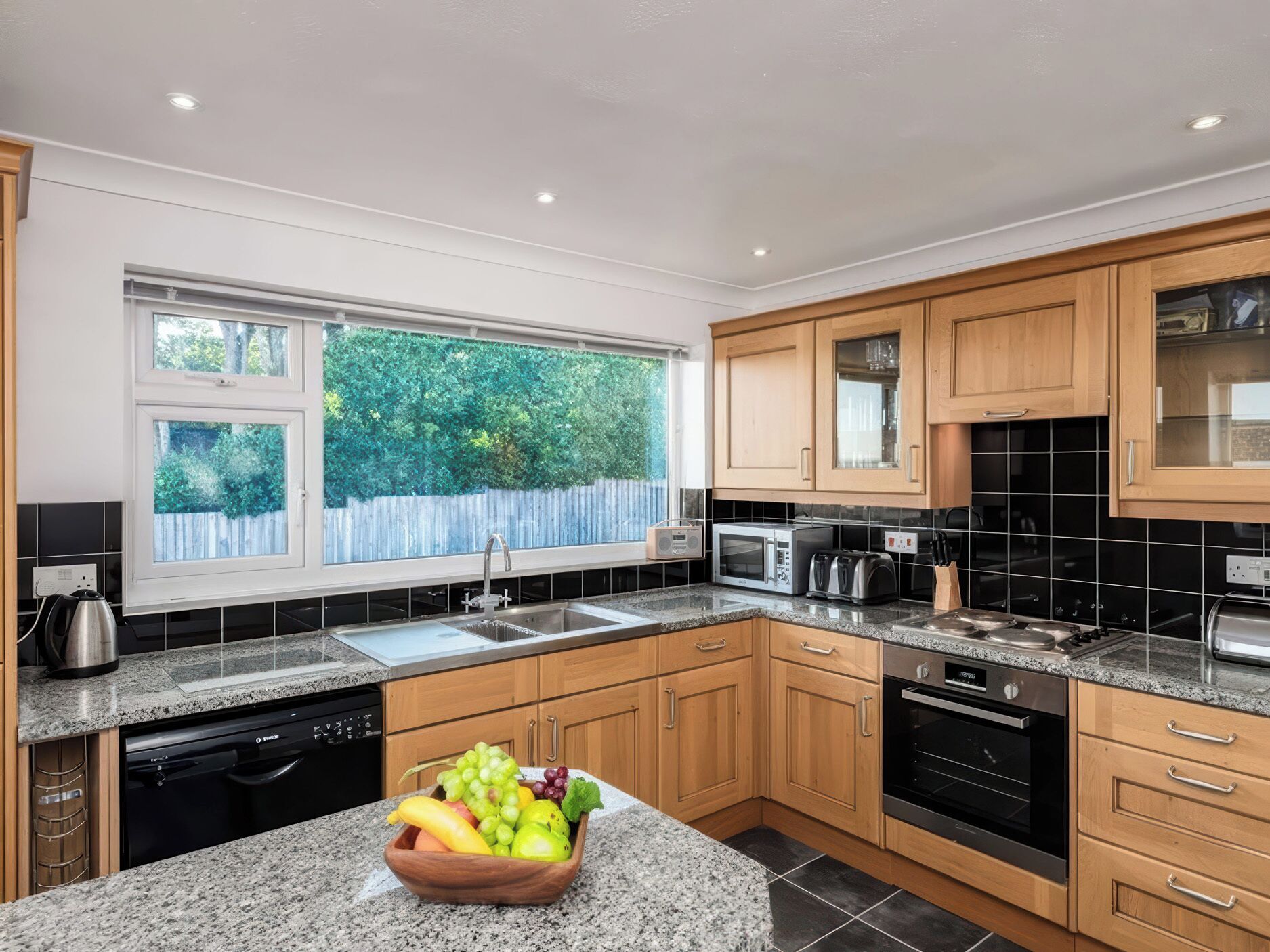 A modern kitchen with wooden cabinets, a granite countertop, a built-in oven, and a window with a view of greenery.