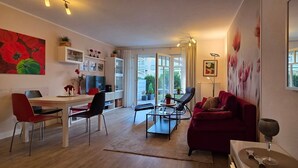 A modern living room with a red sofa, a dining table, and chairs. There is a TV, a bookshelf, and a large window with curtains.