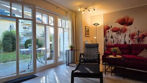 A living room with a red sofa, a black and white checkered chair, a small table with a plant, and a large window with a view of a house and greenery.