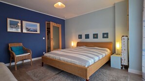 A bedroom with a large bed, a wicker chair, a door, and a nightstand with a lamp.