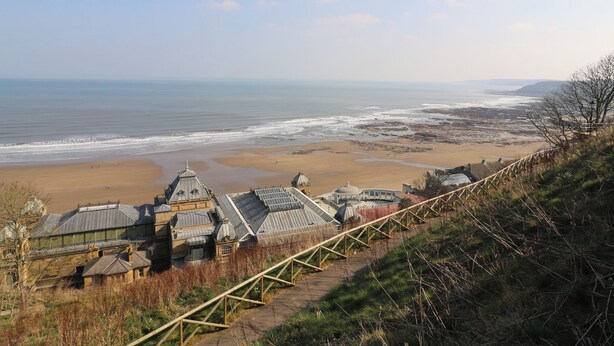A coastal view with a beach, a wooden walkway, and a building with multiple domed structures. - Puffin Cottage (Scarborough)