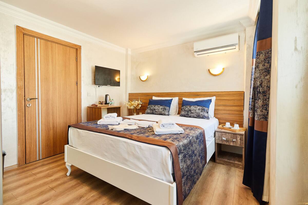 A hotel room with a wooden floor, a bed with a patterned bedspread, a bedside table with a lamp, and an air conditioning unit on the wall.