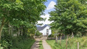 A rural pathway leading to a white house with a wooden gate, surrounded by lush greenery and a mountain in the background. - Drimnatorran Farm Lodge (Acharacle)