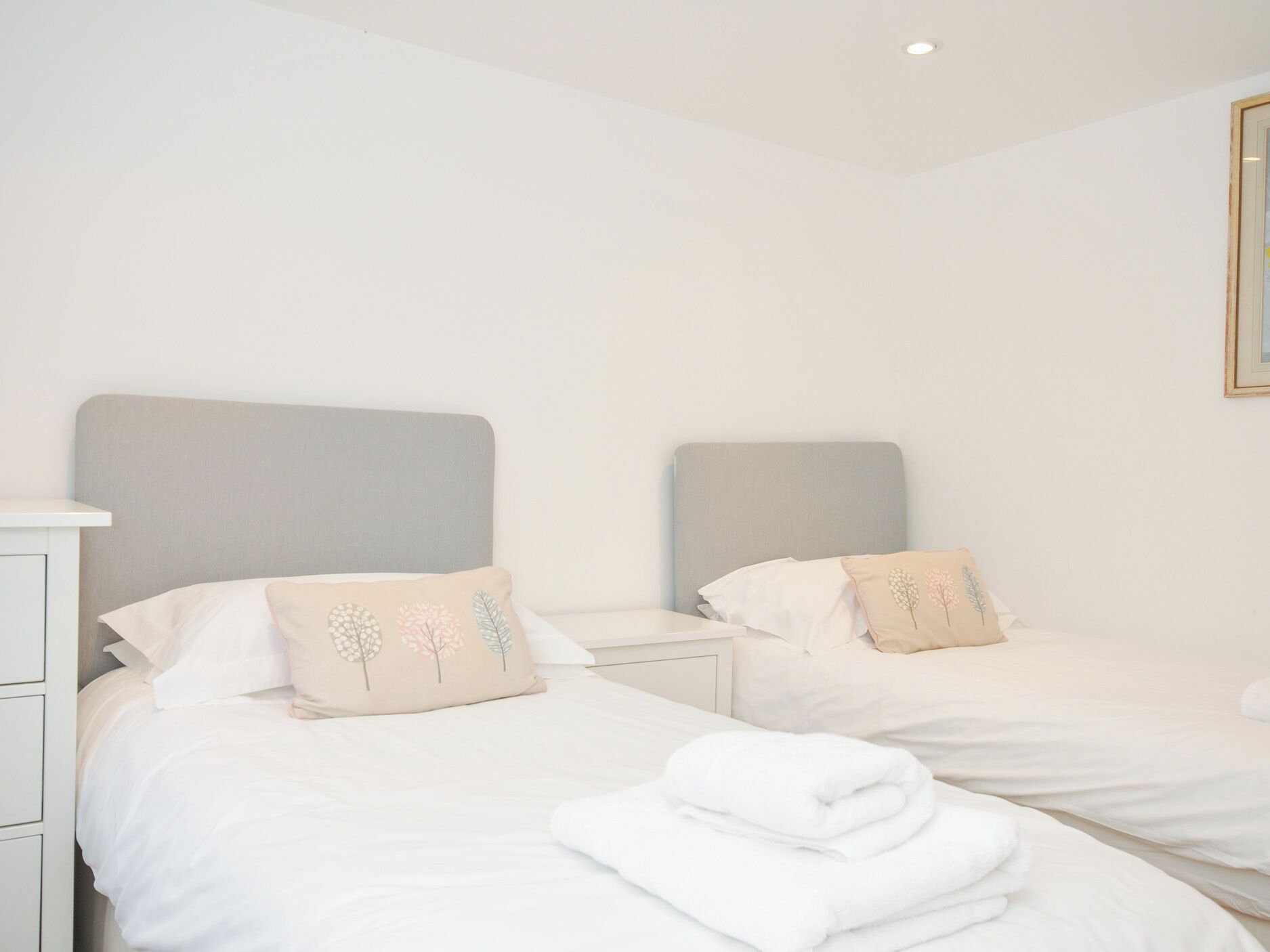 Une chambre d’hôtel avec deux lits simples, chacun doté d’une tête de lit grise et de linge de lit blanc. Il y a une table de chevet blanche sur chaque lit, et un tableau encadré au mur.