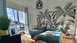 Une chambre avec deux lits, une grande fenêtre, une plante en pot et une fresque murale.