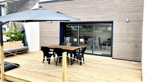 Une terrasse extérieure moderne, avec une table et des chaises en bois, un parasol noir et une porte vitrée donnant sur un espace intérieur.