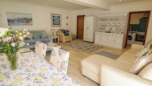 Una sala de estar con sofá, una mesa de centro con mantel floral, un jarrón con flores, un cuadro en la pared y una zona de cocina con lavadora.