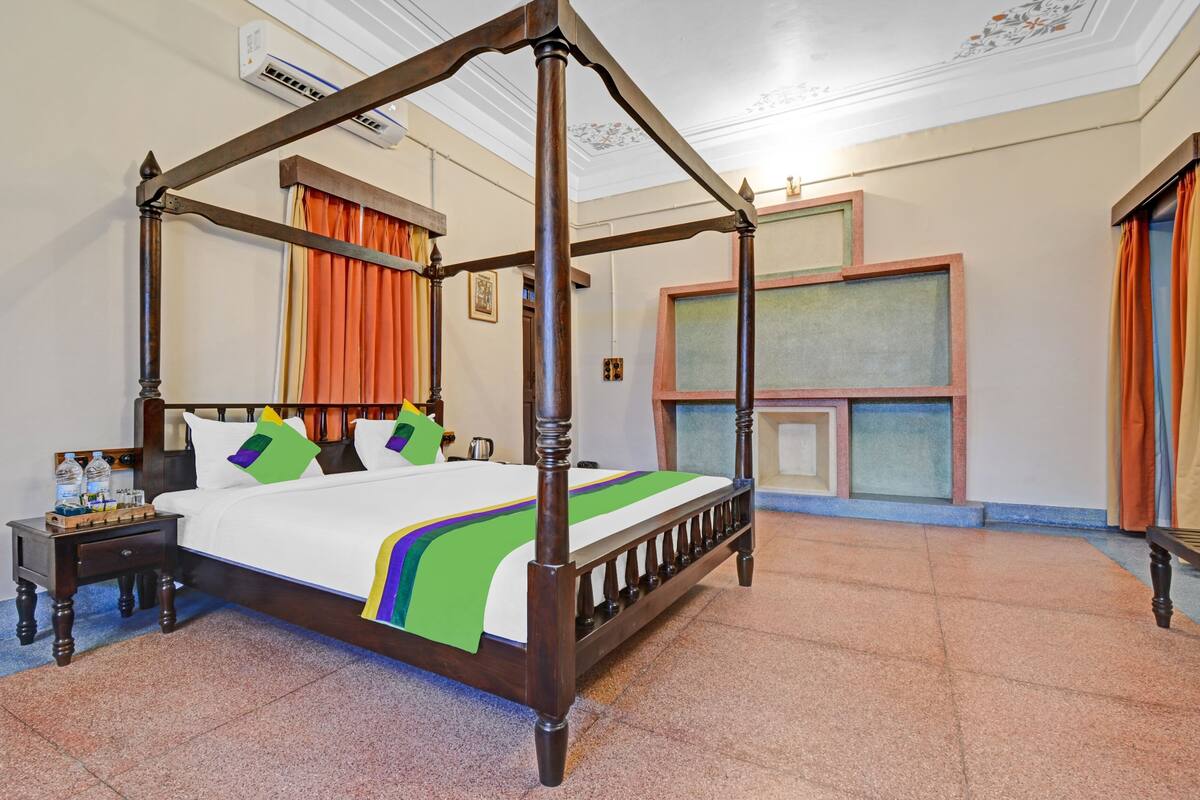 A four-poster bed with a wooden frame, a white bedspread, and colorful pillows. There is a nightstand with a lamp and a water bottle. The room has a tiled floor and a window with orange curtains.