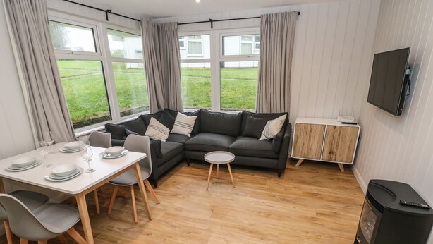 A modern living room with a sectional sofa, a dining table set for four, and a TV mounted on the wall. - Saltwater (Haverfordwest)