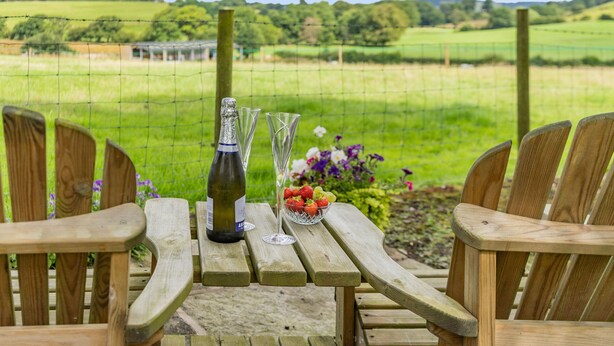 A wooden table with a bottle and glasses, surrounded by chairs, overlooking a green field. - Ingleborough View (Carnforth)