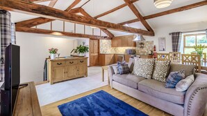 A spacious living room with exposed wooden beams, a sofa with patterned cushions, a television, and a kitchen area in the background. - Ingleborough View (Carnforth)
