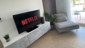 A modern living room with a flat-screen TV on a white console, a grey sofa, a potted plant, and a large window with curtains. - Modern villa with private pool on 5 star Golf Resort (Torre-Pacheco)