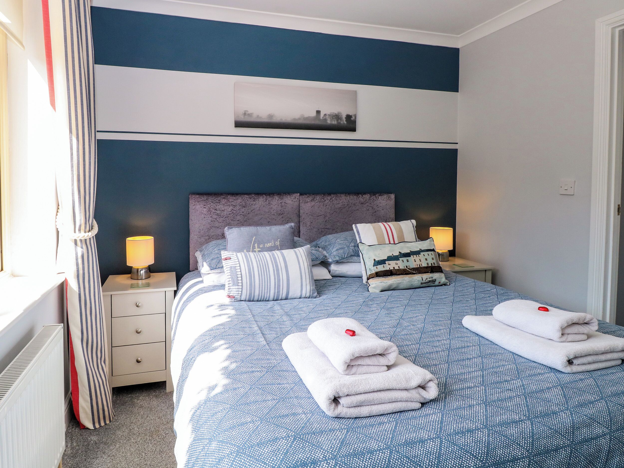 Uma cama bem arrumada com almofadas e uma toalha dobrada, uma mesa de cabeceira com abajur e uma janela com cortinas.