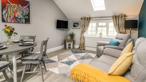 A modern living room with a dining area, grey sofas, a yellow throw, a TV, and a painting on the wall.