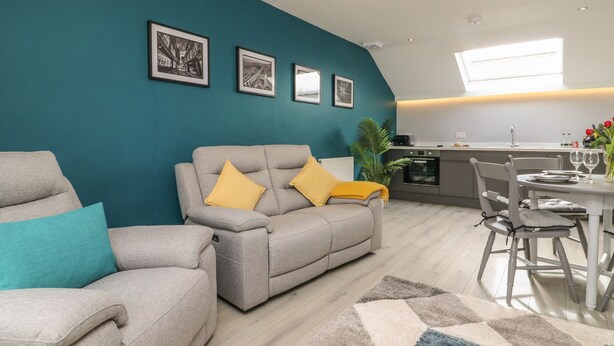 A modern living room with a teal accent wall, grey sofas with yellow cushions, a dining area with a white table, and a kitchen in the background.