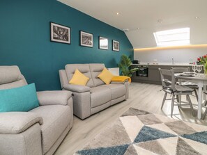A modern living room with a teal accent wall, grey sofas with yellow cushions, a dining area with a white table, and a kitchen in the background. - New Inn Apartment (York)