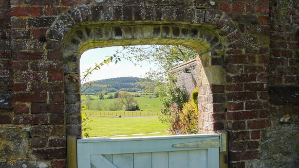 A rustic brick archway with a white wooden gate, leading to a green field and a hill in the distance. - The Stables (Shrewsbury)