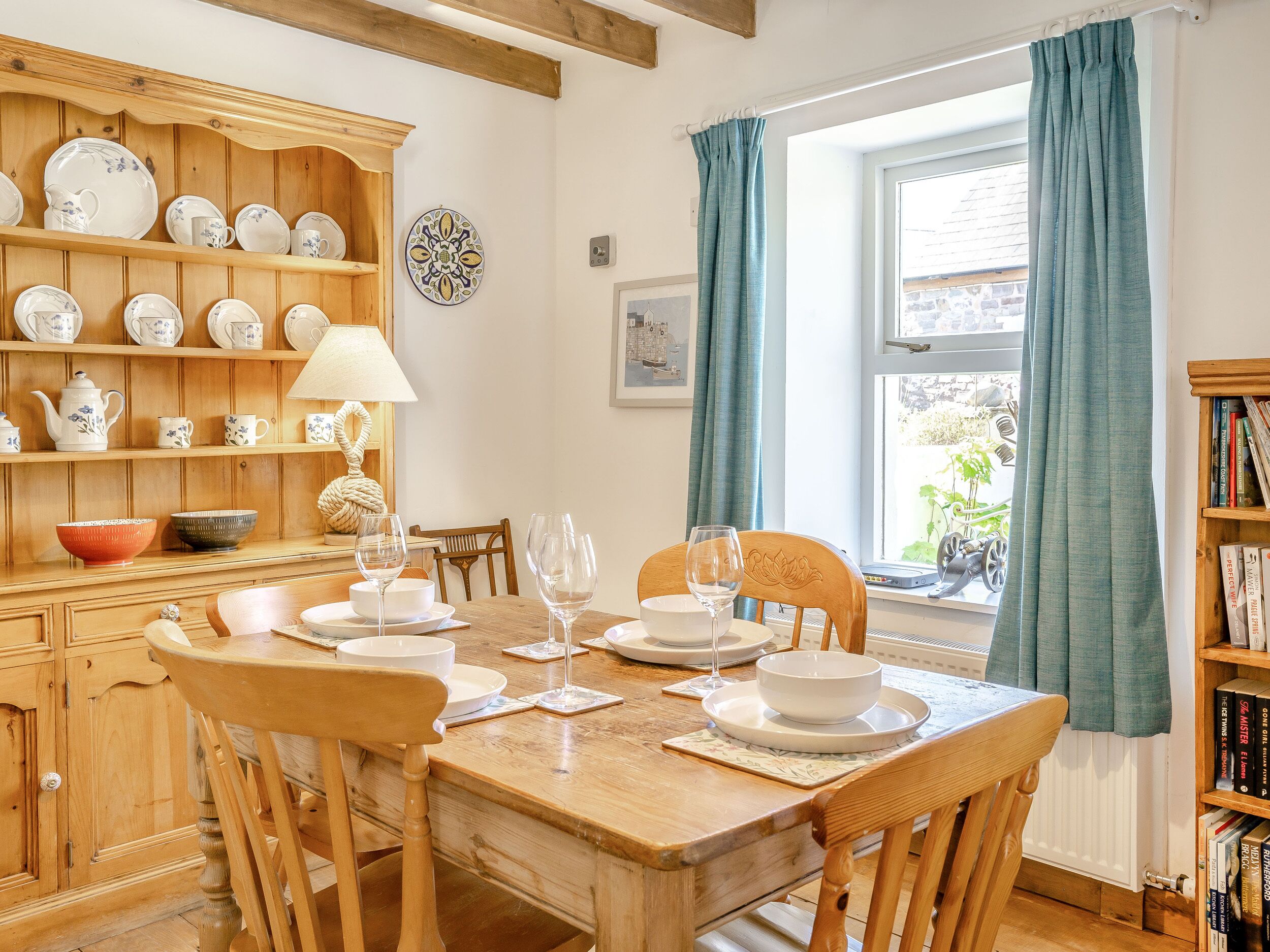 A dining area with a wooden table set for four, surrounded by wooden chairs. A wooden cabinet with dishes is to the left, and a bookshelf is to the right.