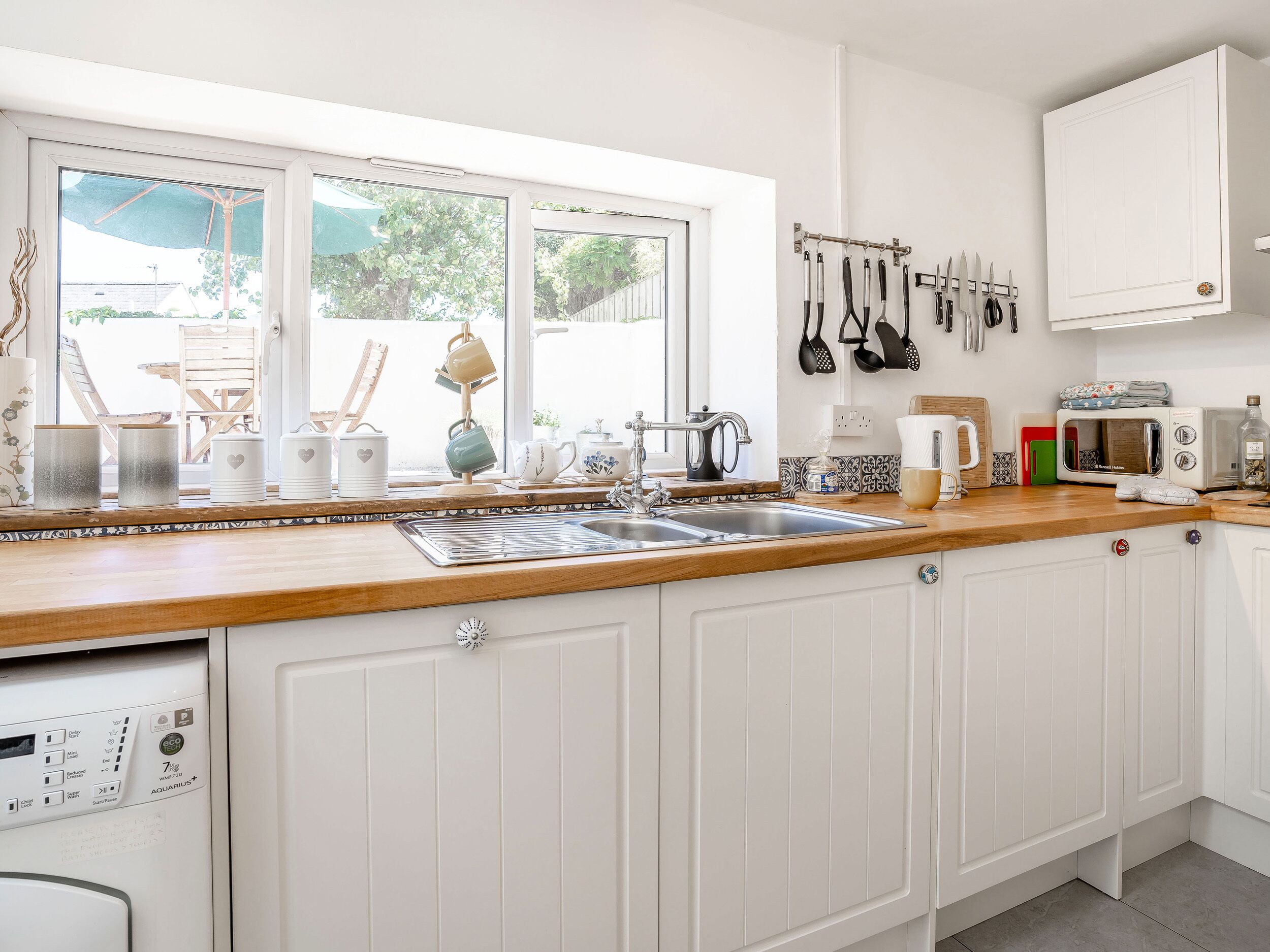 A modern kitchen with white cabinetry, a wooden countertop, a built-in dishwasher, and a window overlooking a garden.