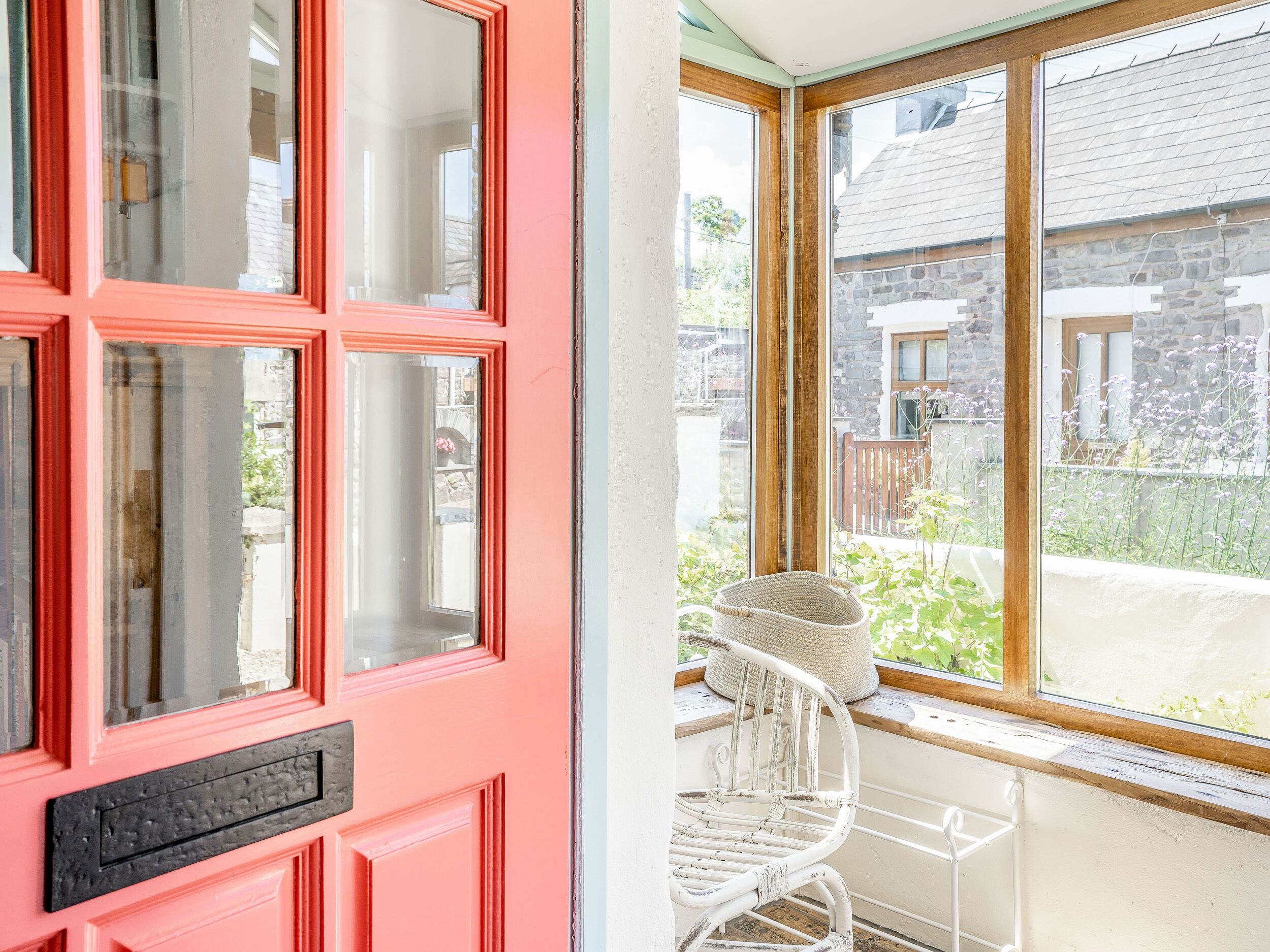 A pink door with multiple glass panes and a white wicker chair by a large window overlooking a garden.