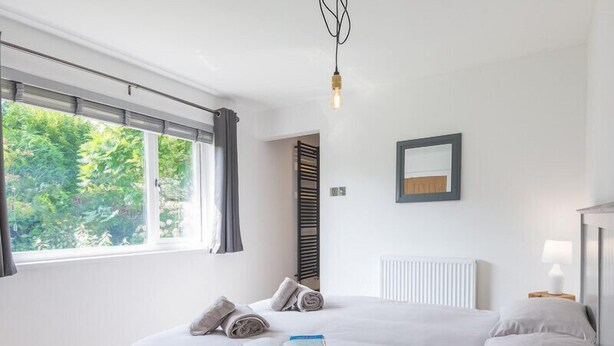 A modern bedroom with a large bed, a view of greenery outside, a radiator, and a bedside table with a lamp. - Greenaway, 4 Rockhaven Gardens (Wadebridge)