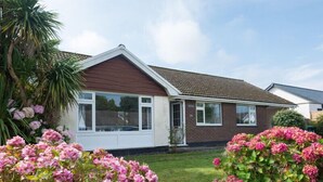 A single-story house with a brown roof and white exterior walls, surrounded by lush greenery and pink flowers. - Greenaway, 4 Rockhaven Gardens (Wadebridge)