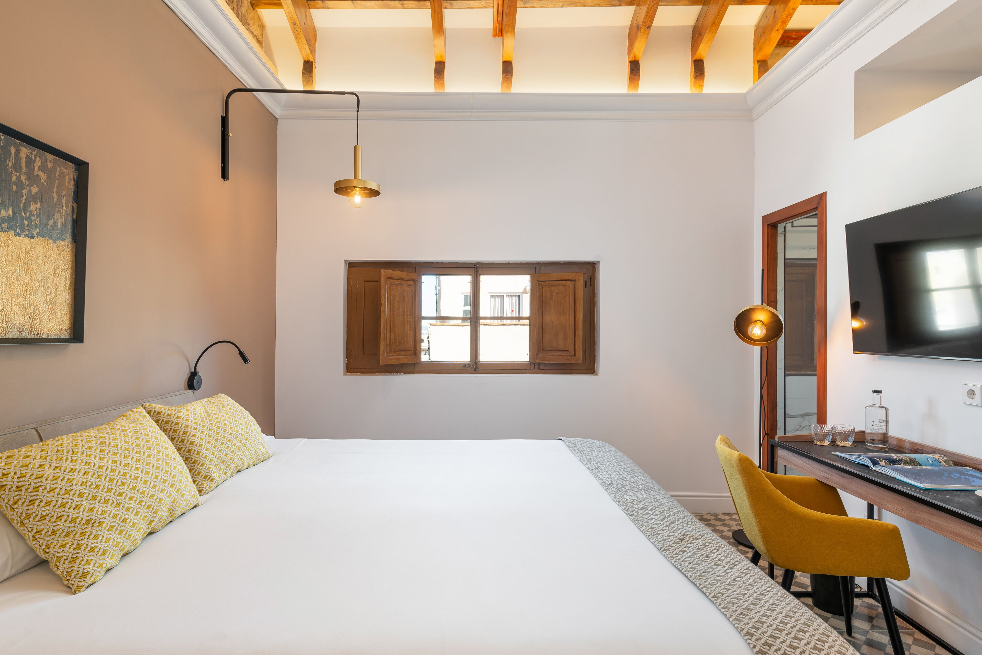 A bedroom with a large bed, a desk with a chair, a window, and a TV mounted on the wall.