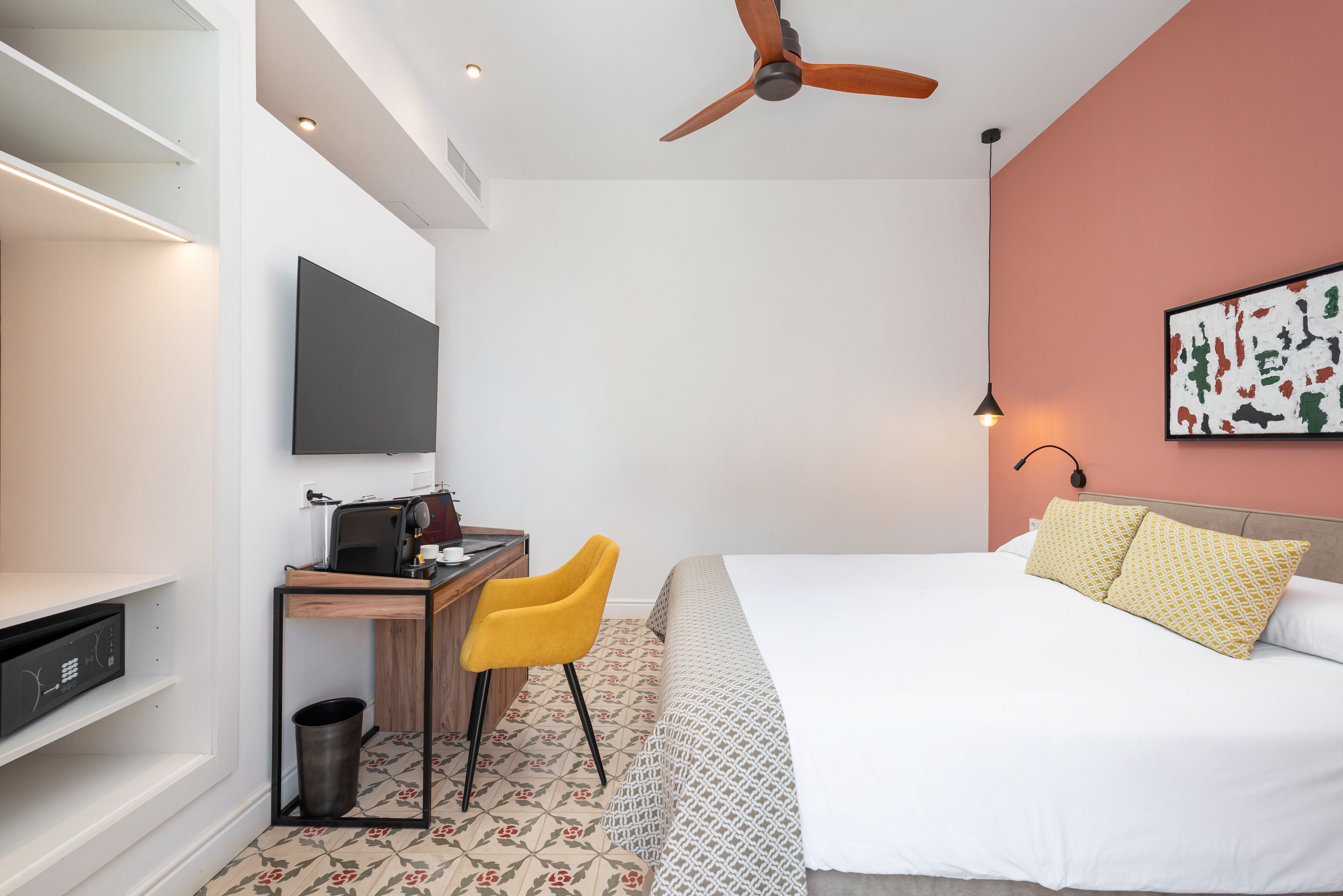 A hotel room with a bed, a desk with a chair, a ceiling fan, a TV mounted on the wall, and a trash can.