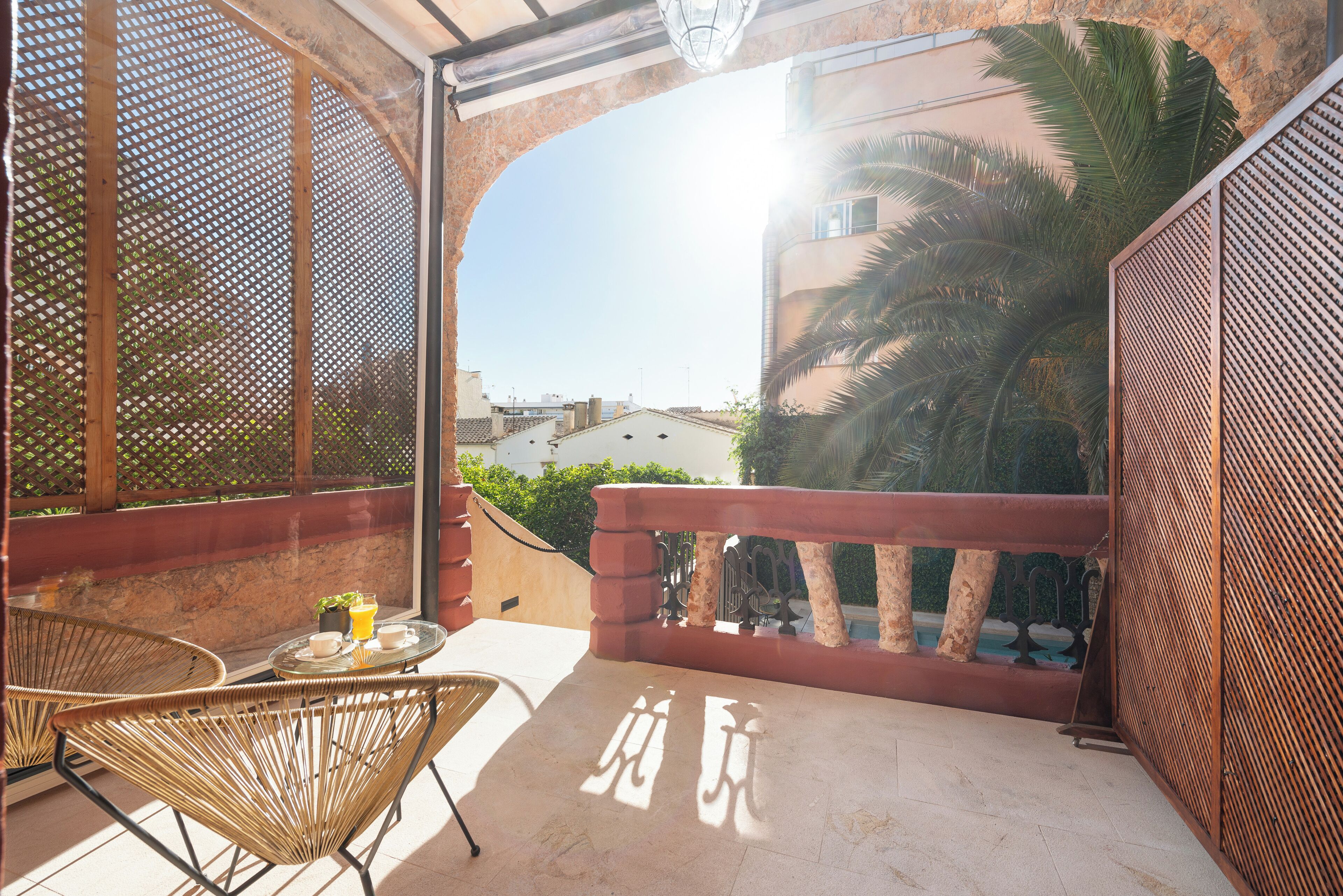 A balcony with a view of buildings and a palm tree, wicker chairs, a table with a tray, and a decorative wall.