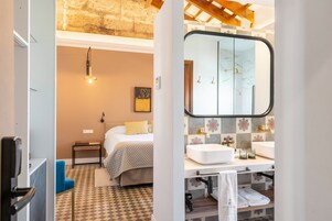 Una habitación de hotel moderna con cama, mesita de noche y baño con un espejo grande, lavamanos y toallero.