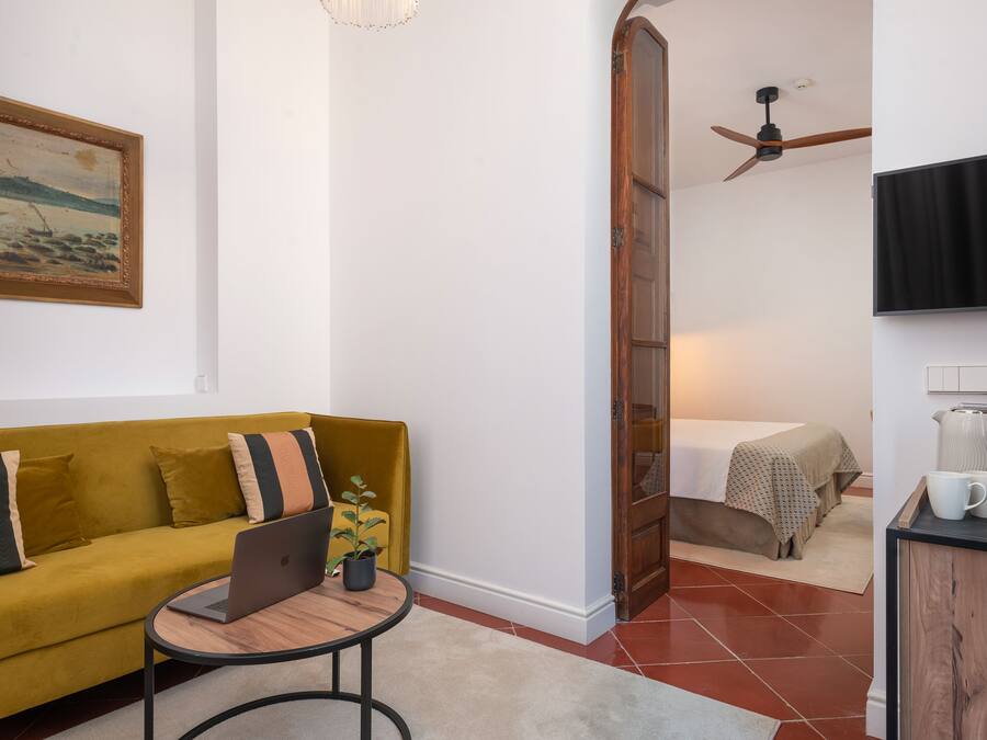A room with a yellow sofa, a laptop on a small table, a ceiling fan, a bed with a white bedspread, and a TV mounted on the wall.