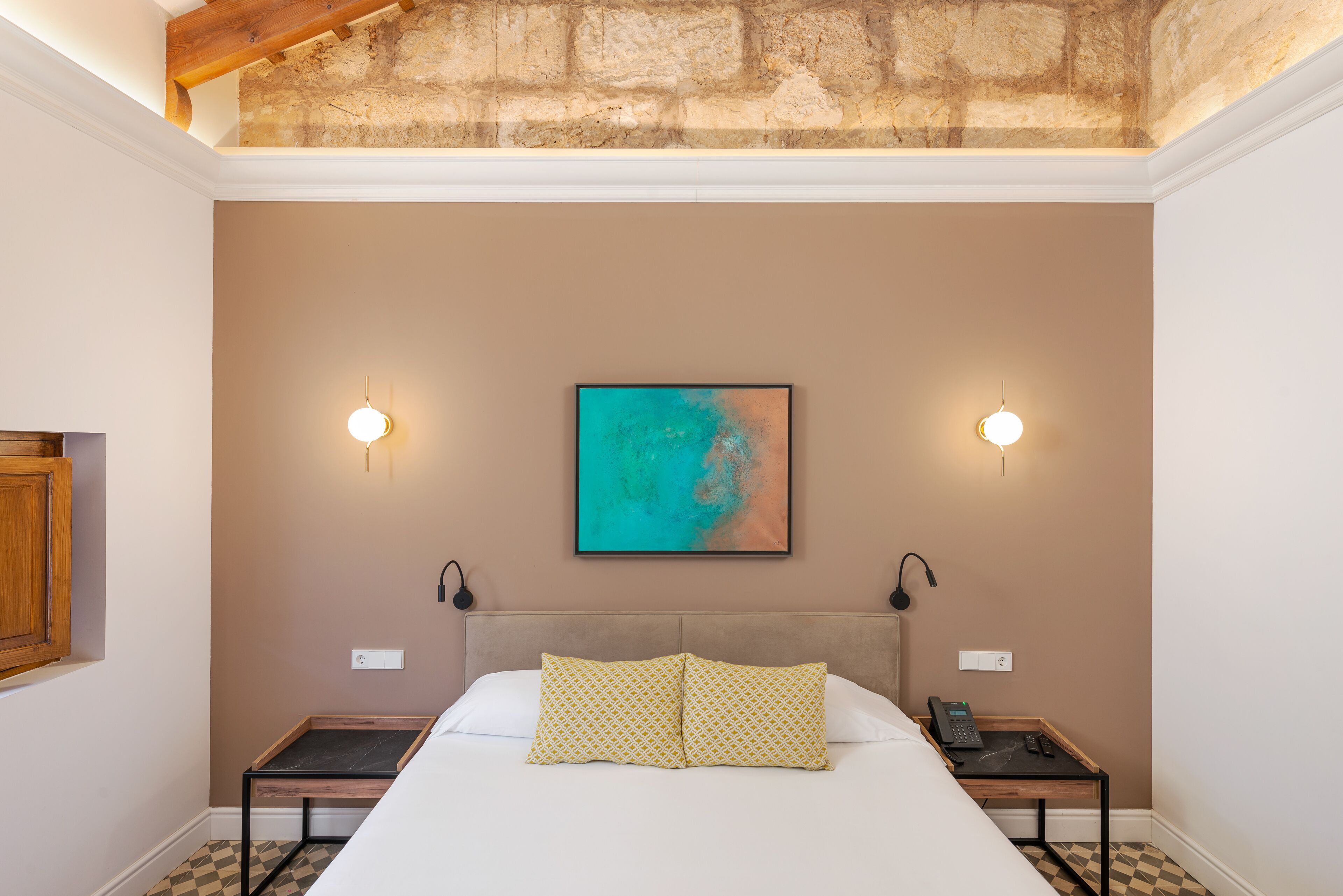 A hotel room with a bed, two bedside tables, a painting on the wall, and a ceiling with exposed beams.