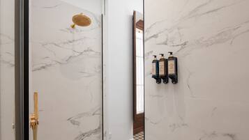 Een badkamer met marmeren wanden, een douche met gouden douchekop en twee flessen shampoo op een zwart schap.