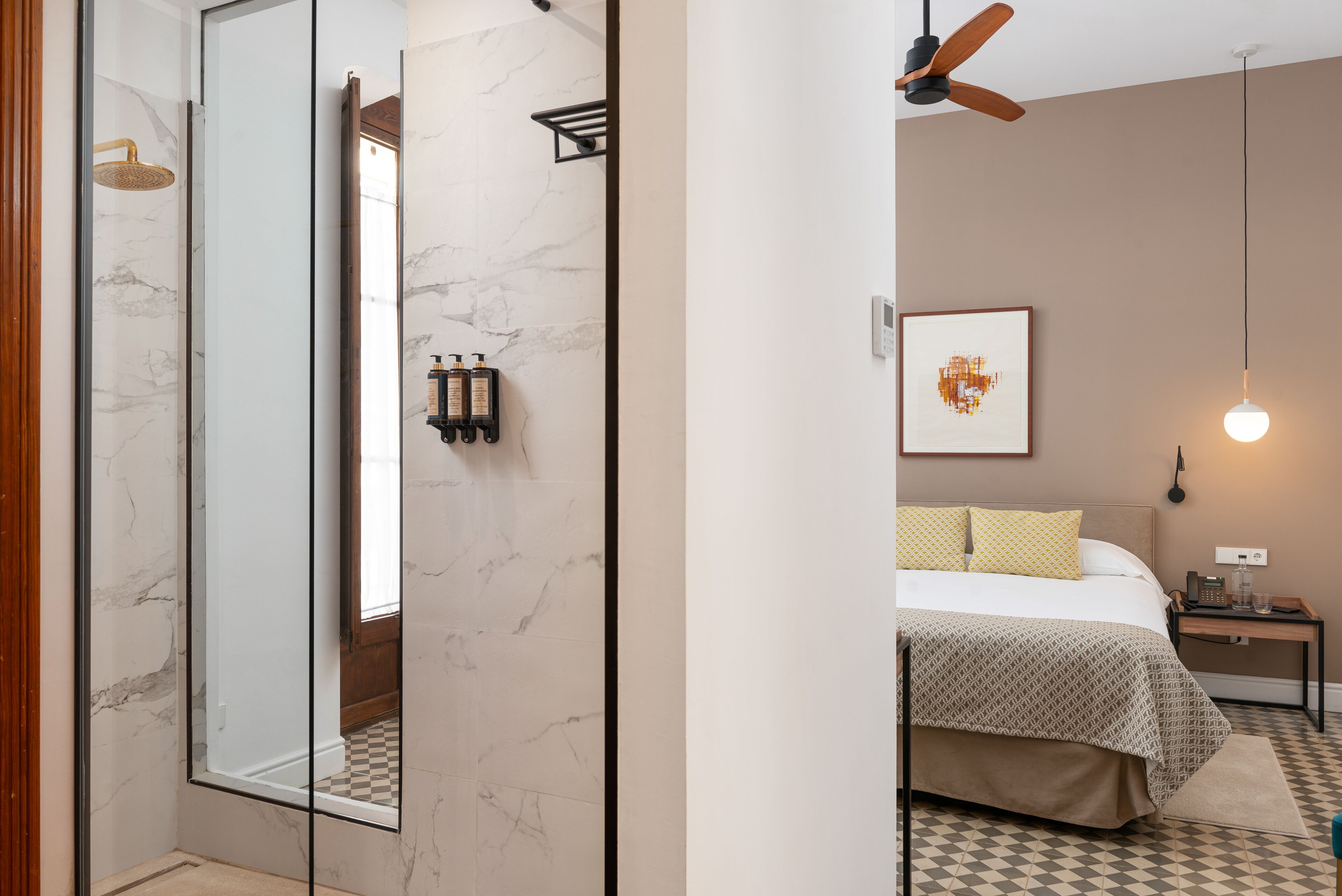 Una habitación de hotel moderna con un espejo grande, una cama con ropa de cama estampada, un ventilador de techo y una obra enmarcada en la pared.