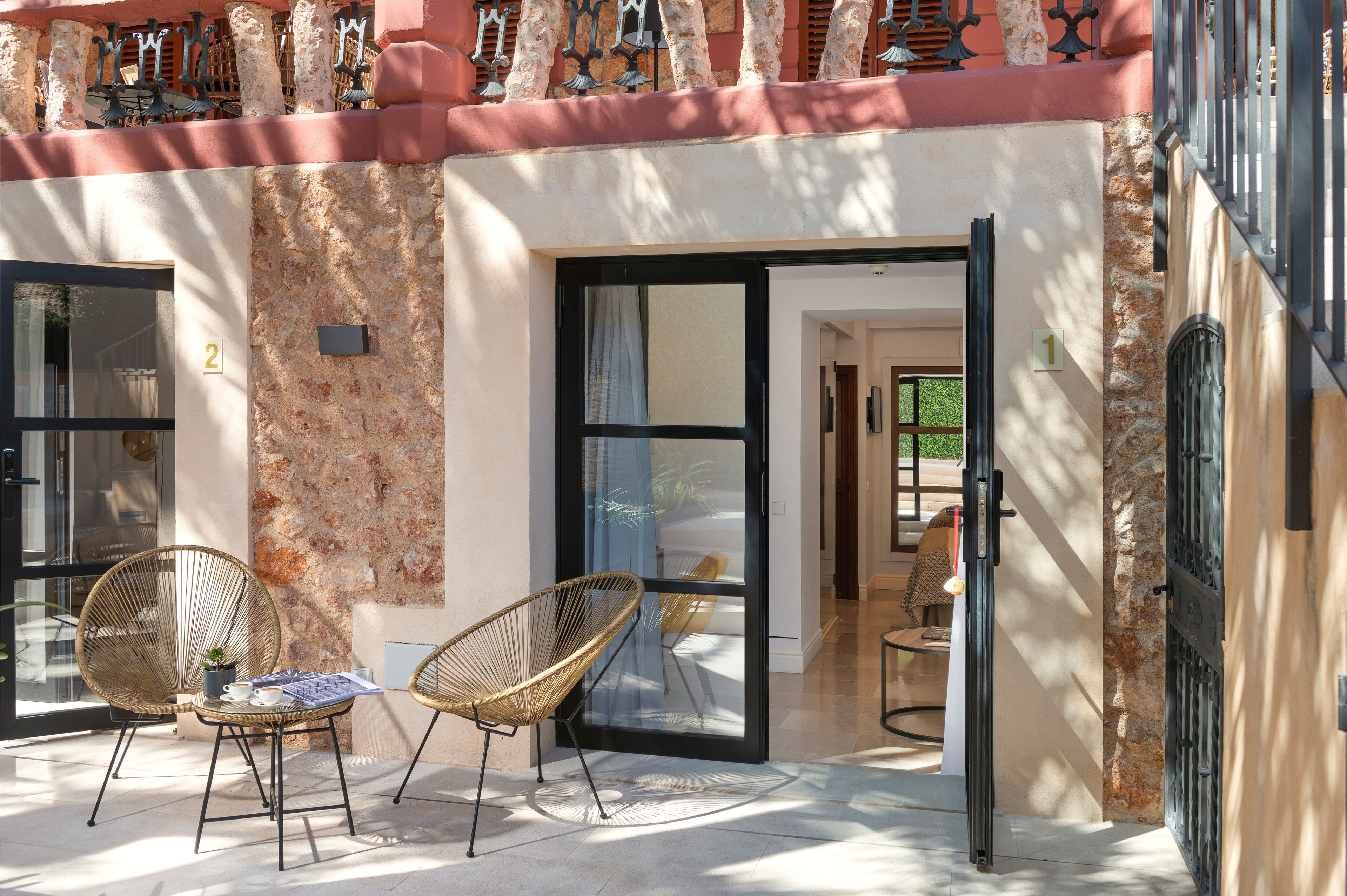 Un espace extérieur aménagé avec des chaises et une table en osier, un mur en pierre et une porte d’entrée en verre.